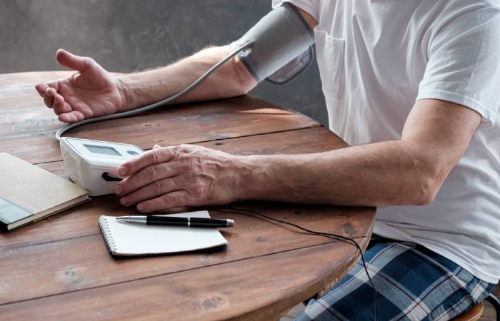 Side view of a man sitting on a chair with a digital BP apparatus cuff wrapped around his right hand, looks into the readings displayed on the monitor along with a pen and notepad kept on a wooden table infront of him.